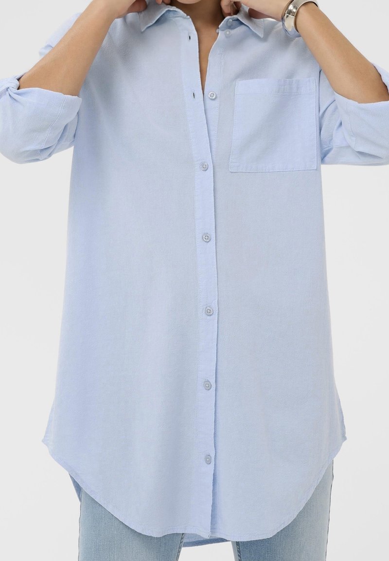 Personne portant une chemise bleu clair à boutons avec manches retroussées et une poche poitrine avant, assortie à un jean bleu clair.