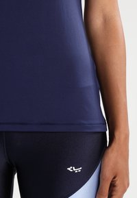 Námořnická sportovní tílko s hladkým povrchem, představující kulatý výstřih a logo na lemu. V kombinaci s tmavě a světle modrými legínami.