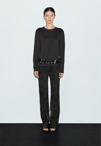 Zwarte lange mouwen top met een leren gevlochten taille detail, gecombineerd met zwarte fluwelen flared broek en open teen muiltjes op een effen achtergrond.