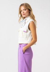 Vit ärmlös crop top med två framfickor och en lila logotagg, tillsammans med vertikalt randiga lavendelfärgade byxor med sidofickor.
