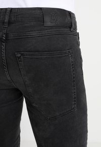 Jeans de mezclilla negra con un ajuste regular, detalle en el bolsillo trasero y tejido texturizado. Muestra un parche de cuero con un logotipo en relieve en la cintura.