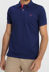 Marinblå polo med röd accentkrage, korta ärmar och en liten röd logotyp på vänster bröst. Tillverkad av texturerad bomull.