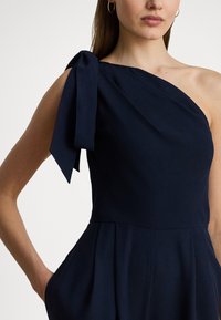 Granatowa sukienka na jedno ramię z dużą kokardą na ramieniu, wykonana z gładkiej tkaniny, z dopasowanym gorsetem i bocznymi kieszeniami.