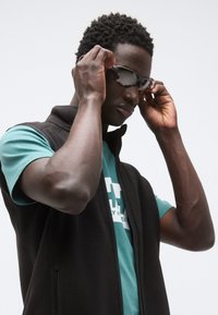 Μαύρο γιλέκο από πολυεστέρα πάνω από ένα πράσινο-μπλε μπλουζάκι, με έναν άντρα που ρυθμίζει τα σκούρα γυαλιά ηλίου του. Έχει κοντά, υφή μαλλιά και βρίσκεται μπροστά σε ένα ανοιχτό φόντο.