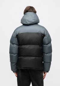 Prešívaná bunda s dvojfarebným dizajnom, kde horná časť je sivá a spodná čierna. Obsahuje kapucňu a elastické manžety. Textúrovaná látka.