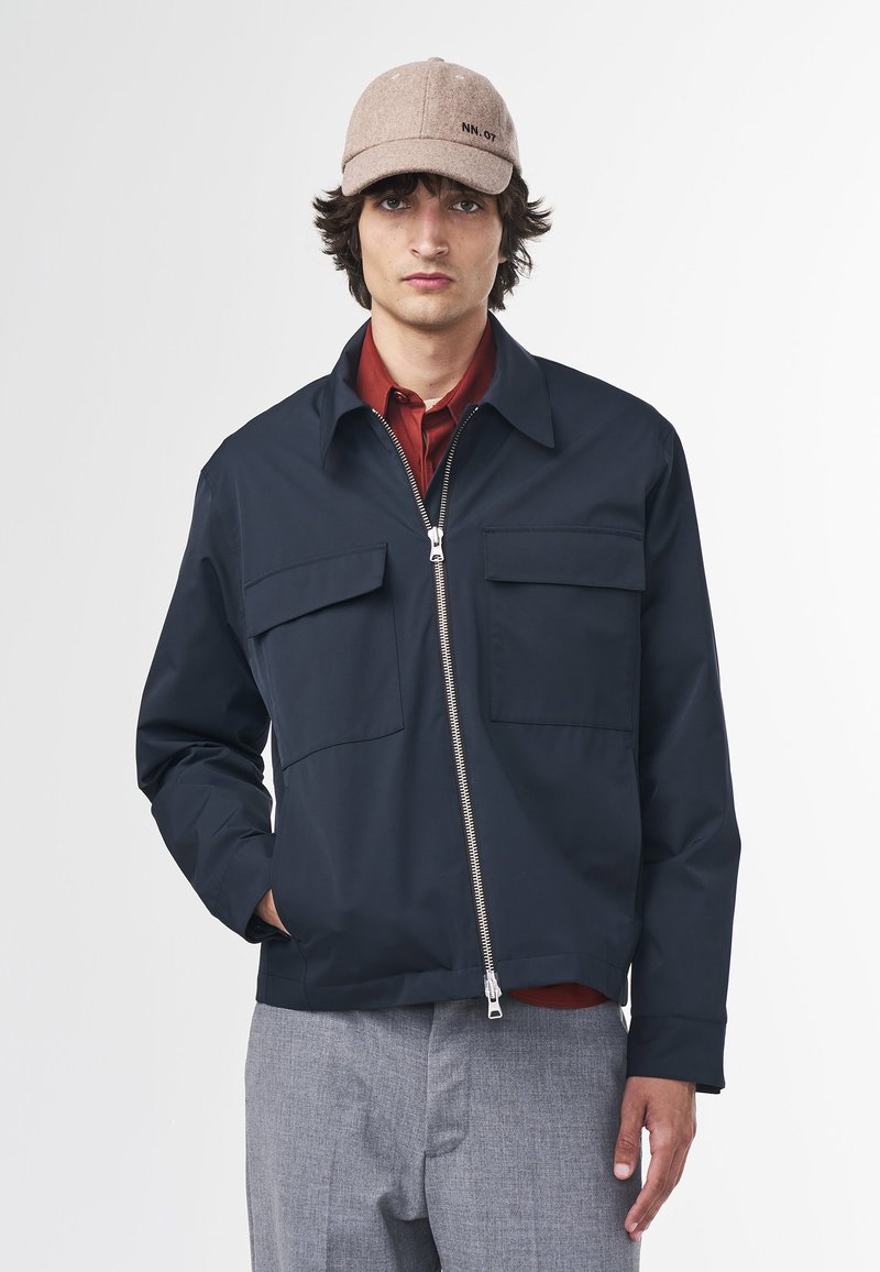 Marinblå zip-up jacka med två bröstfickor, hög krage och en mjuk textur; bärs över en röd skjorta. Ljusgrå byxor fullbordar outfiten.