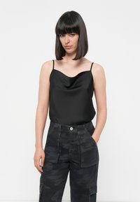 Zwarte satijnen camisole met een cowl-halslijn en dunne bandjes, gecombineerd met donkere camouflage cargobroek met zijzakken en een knoopsluiting.