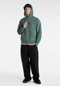 Felpa verde con cappuccio, tasca a marsupio, design a pullover e logo ricamato. Abbinata a pantaloni neri larghi e sneakers nere.
