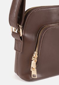 Borsa a tracolla in pelle sintetica marrone con finitura lucida, dotata di chiusura con cerniera dorata e dettagli in metallo. Include una tracolla rimovibile.