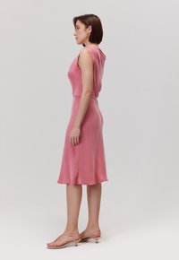 Różowa sukienka bez rękawów, dopasowana w talii, rozszerzająca się na dole. Gładka tekstura z subtelnym połyskiem. Dobrana z jasnymi sandałami.