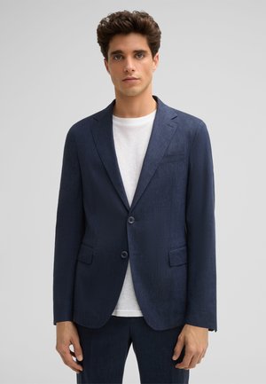 Navy blue, textured blazer with two buttons, notch lapels, and two front pockets. Worn over a white T-shirt, displayed against a light background.