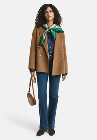 Brauner Doppelreiher-Mantel, goldene Knöpfe, grün-weiße gemusterte Schal, dunkle Denim-Jeans mit Schlag, braune Handtasche, dunkelbraune Schuhe.
