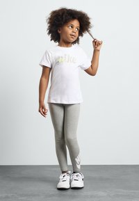 T-shirt branca da Nike com logótipo colorido, leggings cinzentas e sapatilhas preto e branco. Criança de pé com a mão no cabelo, a olhar para cima. Fundo neutro.