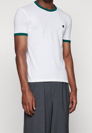 Man draagt een wit T-shirt met korte mouwen en groene bies aan de kraag en mouwen, gecombineerd met grijze geplooide broek.