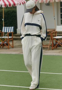 Dviejų dalių apranga: kreminės spalvos užtraukiamas džemperis su tamsiai mėlynomis juostelėmis ir derančios kremo spalvos sportinės kelnės su tamsiai mėlynomis detalėmis, pagamintos iš minkštos medžiagos.