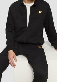 Sudadera negra con cremallera y cuello alto, con un pequeño logo dorado en el pecho; combinada con pantalones deportivos negros a juego con un toque naranja.