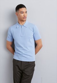 Polo shirt à manches courtes bleu clair avec une fermeture éclair à l'avant, un col et un tissu en maille lisse ; associé à un pantalon sombre.