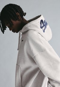 Harmaa huppari, valmistettu pehmeästä kankaasta. Hupussa on kirjailtu tummansininen logo ja rinnassa pieni logo. Mukana kengurutasku.