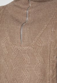 Brauner gestrickter Pullover mit hohem Kragen, teilweise mit silbernem Reißverschluss und strukturiertem Zickzackmuster auf dem Stoff.