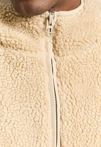 Beige fleecejacka med en texturerad, plyschyta. Har en hel dragkedja och en hög krage. Mjukt material för värme.