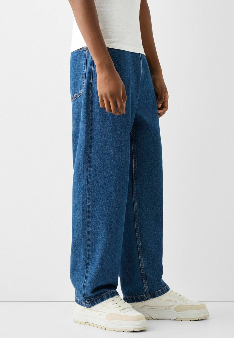 Světle modré oversized džíny s rovnými nohavicemi, s prošíváním kapes a kontrastním šitím, v kombinaci s bílými teniskami.