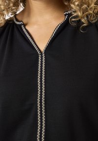 Czarna bluzka z dekoltem w serek, ozdobiona srebrnym przeszyciem wzdłuż listwy guzikowej. Gładka tkanina o miękkiej fakturze.