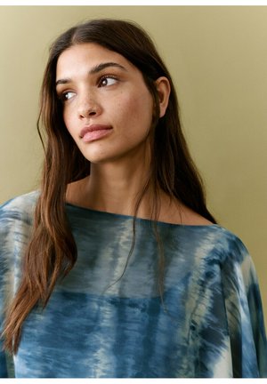 Tânără cu păr lung și castaniu, purtând un top albastru cu imprimeu tie-dye, privindu-se gânditoare într-o parte, pe un fundal simplu bej.