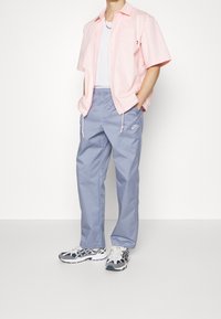 Camicia a maniche corte di colore rosa chiaro con bottoni, canotta bianca, pantaloni leggeri blu con cordoncino e sneaker con motivo. Logo Nike ben visibile.
