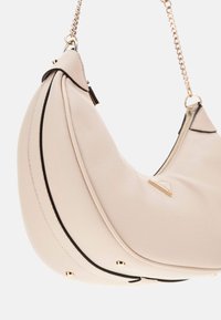 Sac à main en cuir beige avec une forme de croissant, piping noir, quincaillerie dorée et une bandoulière en chaîne. Dispose d'une fermeture éclair et d'éclats décoratifs.