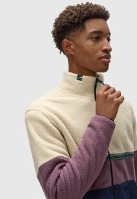 Chaqueta de forro polar en crema, morado y azul marino. Cuenta con un alto cuello, media cremallera, tela texturizada y un detalle de rayas estampadas en el pecho.