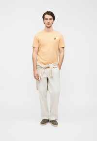 Πορτοκαλί βαμβακερό μπλουζάκι με μικρό μαύρο λογότυπο, σε συνδυασμό με ανοιχτό μπεζ παντελόνια με φαρδιά πατζάκια και πράσινα παπούτσια, με μπεζ πουλόβερ δεμένο στη μέση.