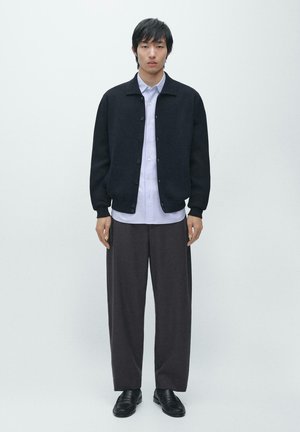 Veste noire boutonnée, chemise blanche et pantalon gris à jambes larges. Tissu lisse, design structuré et style minimaliste. Chaussures noires.