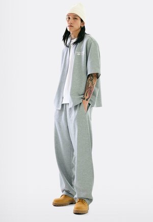 Graues Strickshirt mit kurzen Ärmeln und Knöpfen, kombiniert mit lockeren grauen Sweatpants. Das Modell trägt ein weißes T-Shirt darunter und beigefarbene Stiefel.