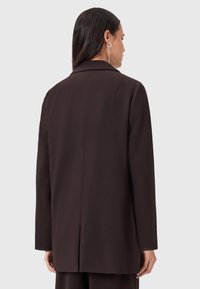 Blazer marrone su misura con colletto a rever, schiena dritta e maniche lunghe. Texture liscia, dettagli minimi e linee pulite.