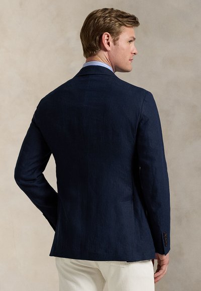 Tengerészkék lenblézer szabott fazonban. Egyetlen hátsó sliccel, beszabott hajtókával és gombos mandzsettával rendelkezik. Világos színű nadrág felett viselve.