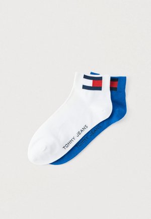 Δύο ζευγάρια κάλτσες αστραγάλου: ένα λευκό με μπλε και κόκκινη λωρίδα λογότυπου, και ένα ολόκληρο μπλε με την επιγραφή "TOMMY JEANS" σε λευκό.