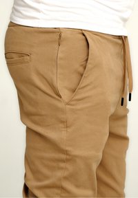 Beige Jogginghosen aus weichem Stoff, mit einer seitlichen Reißverschlusstasche, einem Kordelzugbund und einer glatten Oberfläche.