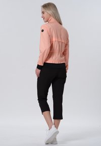 Kevyt persikanvärinen takki, jossa on korkea kaulus, elastisoitu helma ja mustat hihansuut, yhdistettynä mustiin lyhytlahkeisiin housuihin ja valkoisiin lenkkareihin.