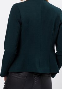 Blazer ajusté en tissu texturé couleur bleu sarcelle foncé, avec une coupe cintrée et des coutures aux épaules, porté avec un pantalon noir en similicuir.
