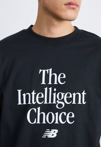 Musta puuvillane T-särk, millel on suur valge tekst "Intelligentne valik" ja New Balance'i logo, lühikesed varrukad ja ümar kaelus.