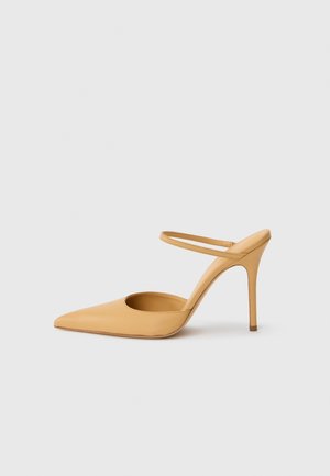 Beige, hegyes orrú stiletto sarokcipő, vékony bokapánttal, fehér háttér előtt.