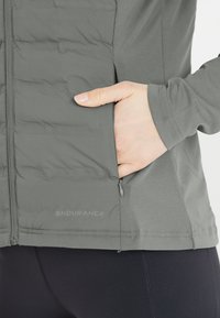 Chaqueta ligera gris con textura acolchada, bolsillos con cremallera y dobladillo elástico. Incluye la marca 'ENDURANCE'.