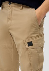 Lichte kaki cargobroek met een gladde textuur, zijzak en een zwart logo-embleem. Ontworpen met een traditionele rits en knoopsluiting.