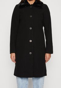 Manteau noir avec un col en fausse fourrure, fermeture à boutons, manches longues et poches latérales ; fabriqué en tissu texturé avec une coupe droite.