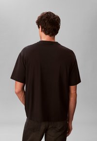 Άνδρας με κοντά καστανά μαλλιά, φοράει απλό μαύρο κοντομάνικο μπλουζάκι και σκούρο τζιν, φαίνεται από πίσω με γκρι φόντο.