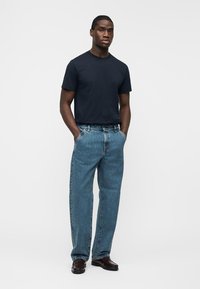 Mand stående med hænder i lommerne iført en sort t-shirt, løse blå jeans og mørkebrune sko mod en hvid baggrund.