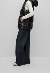 Svart pufferväst med huva, med en quiltad design och framdragkedja, bärs över en långärmad vit topp och mörk vida jeans.