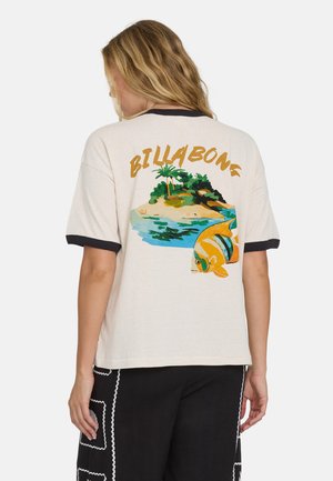 Kobieta zwrócona tyłem, ubrana w beżowy T-shirt z czarnym obszyciem, z grafiką tropikalnej wyspy i ryb oraz napisem "BILLABONG" na plecach.