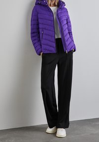 Fioletowa kurtka puchowa z kapturem, z poziomym pikowaniem. W połączeniu z czarnymi spodniami o szerokich nogawkach i białymi trampkami.