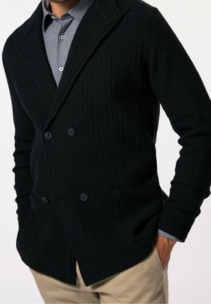 Uomo che indossa un cardigan nero strutturato doppiopetto sopra una camicia blu a fantasia con bottoni e pantaloni beige.